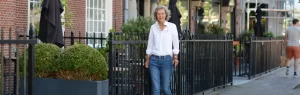 Een vrouw van middelbare leeftijd loopt met een wandelstok over de Kneuterdijk in den Haag. Ze loop langs het hek van een prachtig oud pand. Het is mooi weer en zij draagt een witte blouse en blauwe jeans. Zij kijkt lachend in de lens.